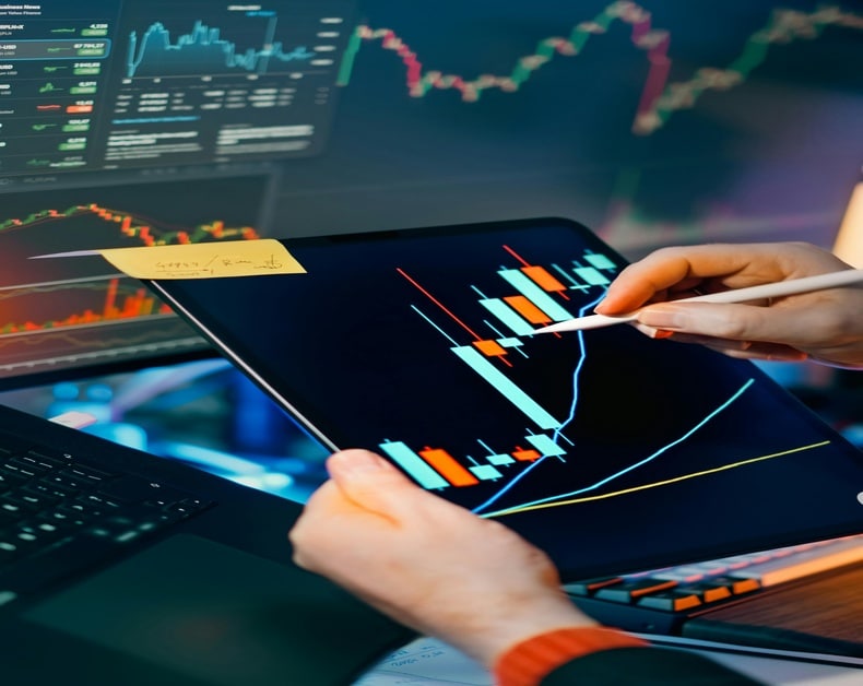 Analyst using a stylus to review candlestick charts on a tablet beside a laptop showing market graphs.