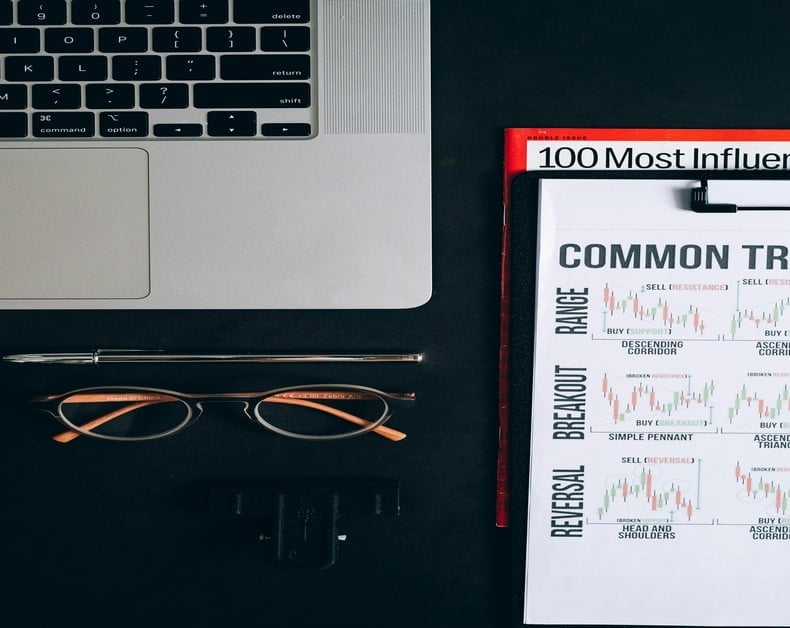 Top-down desk scene with a silver laptop, glasses, and a pen on a dark surface beside a clipboard of charts labeled 'COMMON' and '100 Most Influ...'.