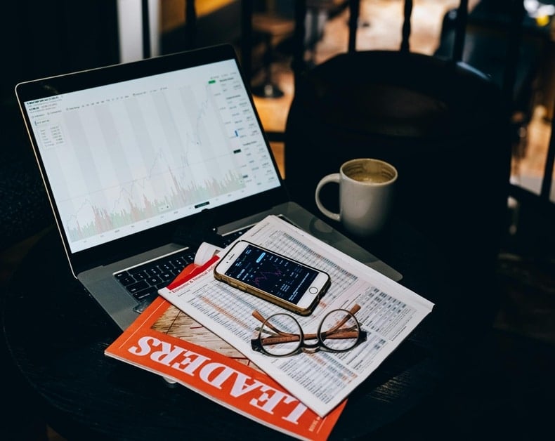Laptop shows stock charts on a cafe table, with a smartphone displaying market data, a coffee mug, newspaper, and glasses nearby.
