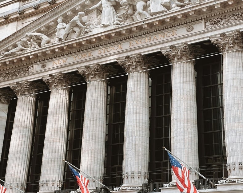 Front of the New York Stock Exchange with tall columns and neoclassical frieze, US flags fluttering below.