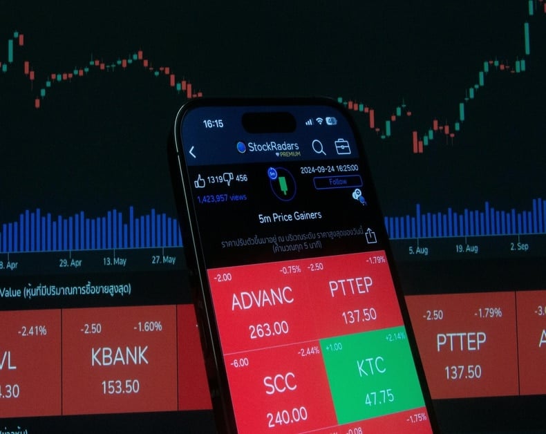 Close-up of a smartphone displaying a stock trading app with red/green price tiles and a dark market chart in the background.