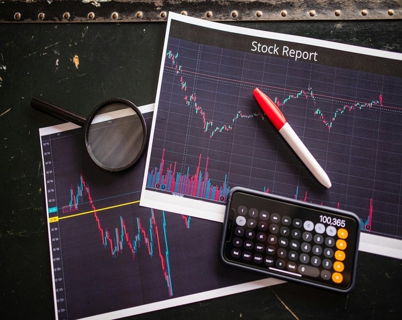 Stock charts and a calculator on a dark desk, with a magnifying glass and red-marked pen nearby.