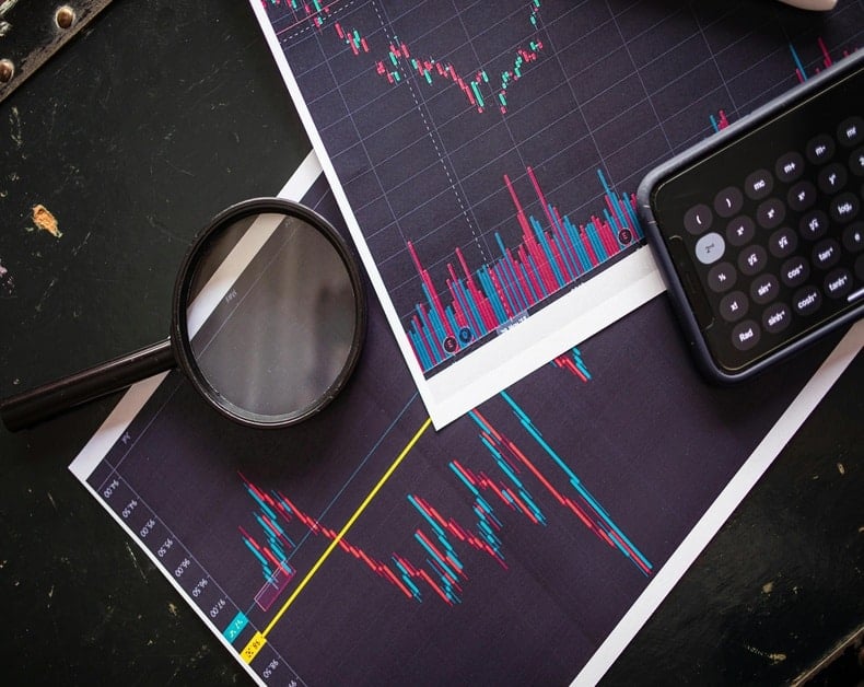 Stock market charts with red and blue price bars, a magnifying glass, and a calculator on a dark desk