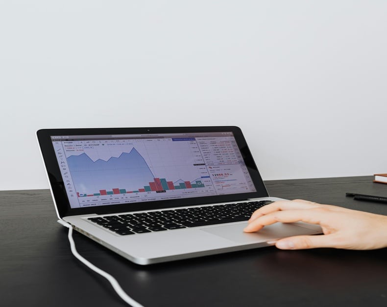 Laptop on a dark desk displaying a financial chart with blue line and colorful bars; a hand rests on the trackpad.