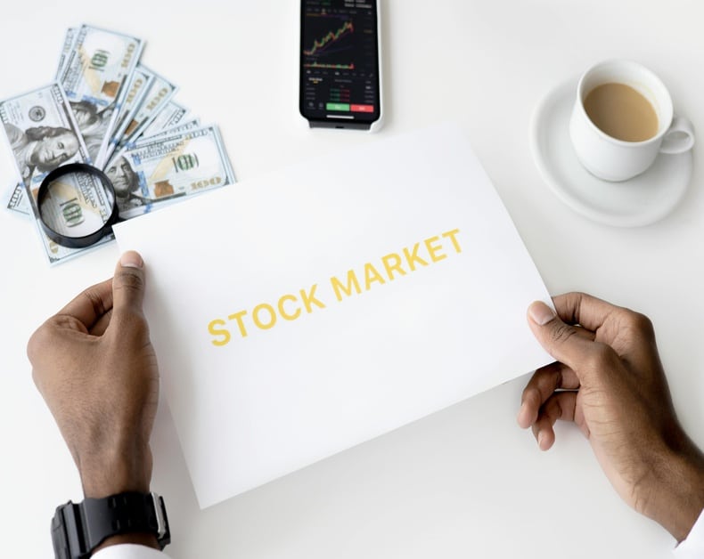 Hands hold a white sheet labeled STOCK MARKET on a desk with cash, a magnifying glass, a charting phone, and a cup of coffee.