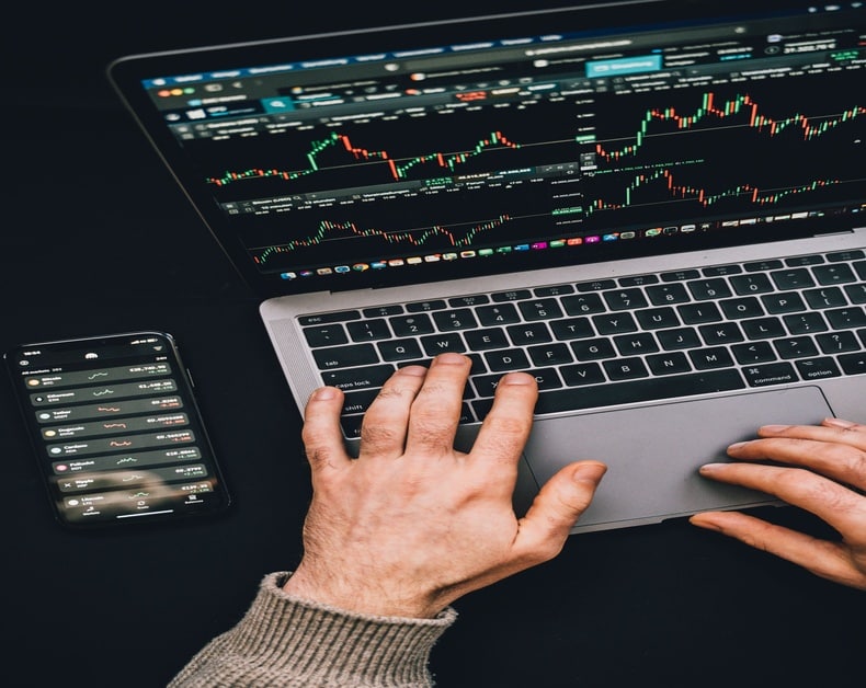 Hands typing on a laptop displaying stock charts, with a smartphone showing market data beside it.