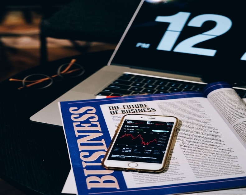 Desk scene with a business magazine titled 'The Future of Business', a smartphone displaying stock charts resting on the magazine, a laptop showing a large '12' on screen, and a pair of glasses nearby