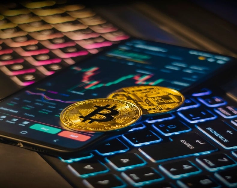 Gold Bitcoin coins rest on a smartphone showing a crypto trading app with buy/sell buttons and market charts, on a backlit keyboard.