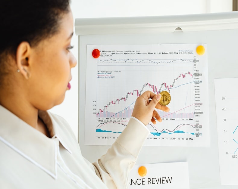 Person holding a gold Bitcoin coin over a trading chart on a whiteboard, suggesting cryptocurrency investment or market analysis.