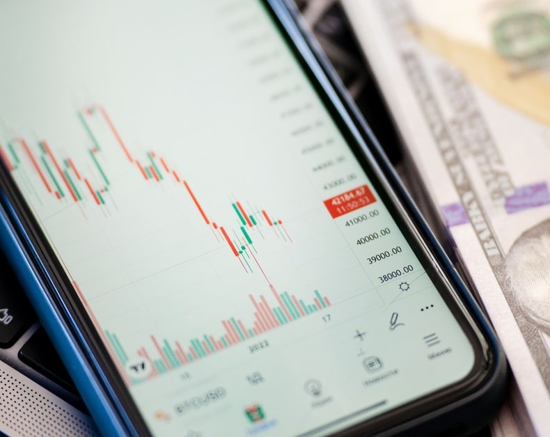 Smartphone screen showing a red and green candlestick stock chart beside folded US dollars.