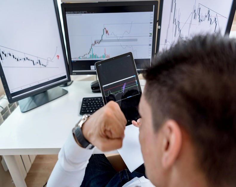 Trader fist-pumping while monitoring stock charts on a tablet and multiple monitors in the background.