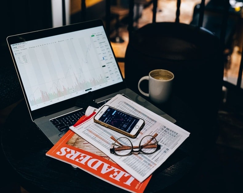 Stock trading workspace: laptop showing market charts, smartphone with data, glasses, a newspaper, and a coffee mug on a dark desk beneath soft lighting.