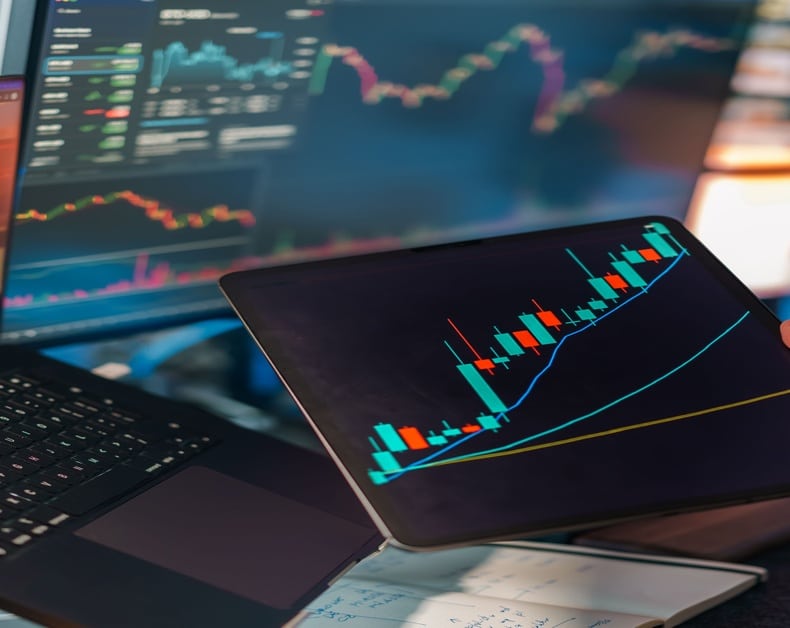 Tablet showing a colorful candlestick stock chart with an upward trend, with a laptop and multiple market dashboards in the background