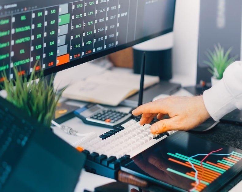 Close-up of a hand typing on a white keyboard while stock charts glow on a large monitor behind it, at a finance desk.