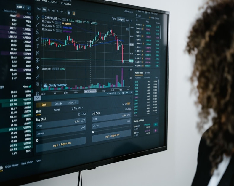 Trading dashboard on a monitor showing candlestick chart, volume bars, and market data for CAKESLT on screen, with a person in the background.