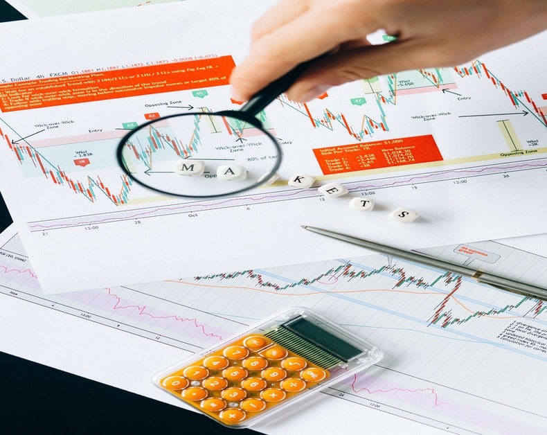 Person analyzing stock charts with a magnifying glass, calculator, and notes on a desk.