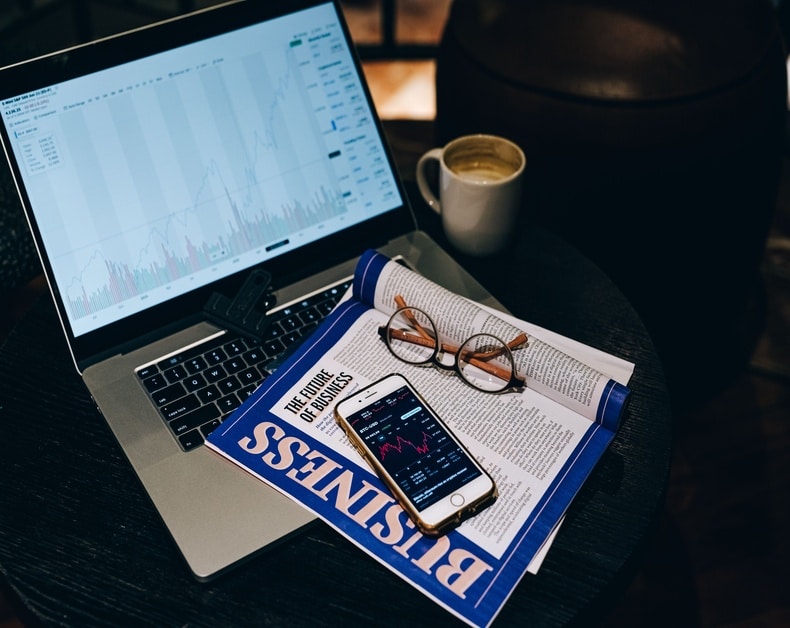 Laptop with stock charts open, a Business magazine, reading glasses, a smartphone displaying market data, and a mug of coffee on a dark table.