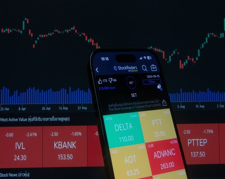 Close-up of a smartphone showing a color-coded stock trading app over a dark trading dashboard with charts in the background.