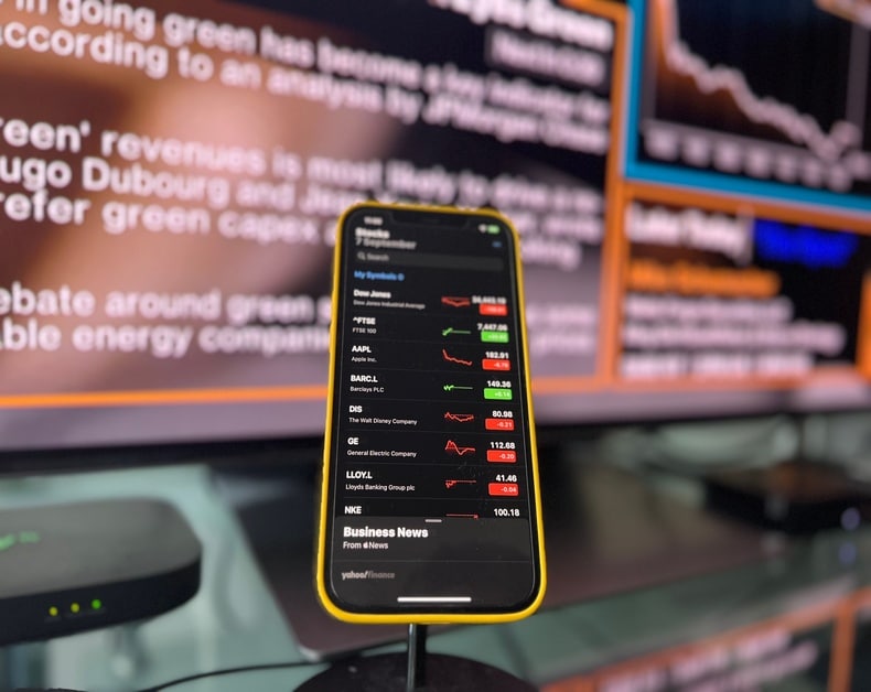 Phone in a yellow case shows a stock market app with red/green price changes, foreground with blurred monitors behind.