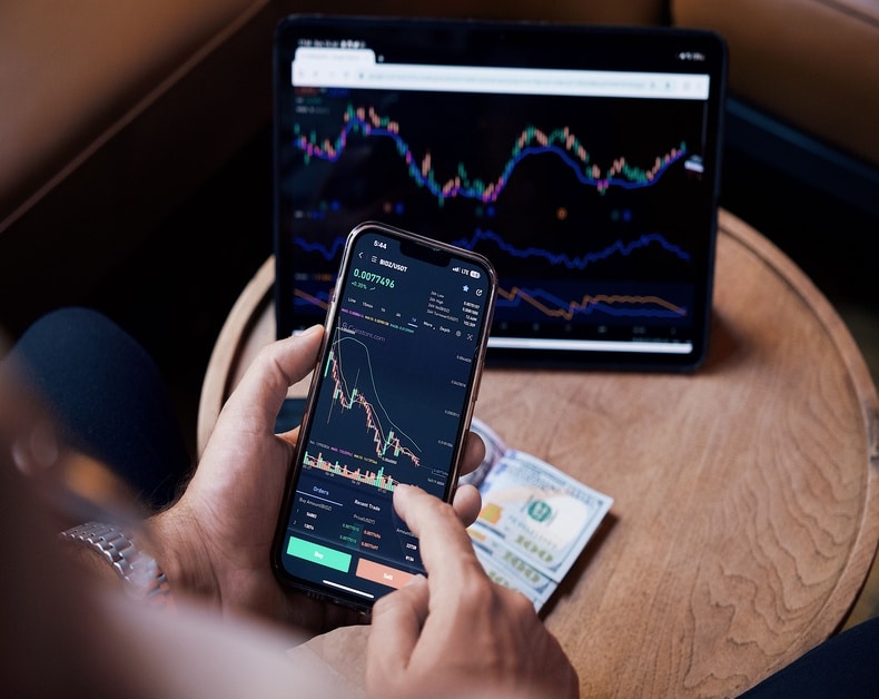 Person holds a smartphone with a trading app showing candlestick charts and buy/sell controls; a tablet with market graphs sits on a round wooden table with cash nearby.