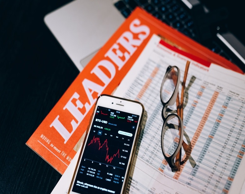 Smartphone showing a red stock chart on a desk with an orange LEADERS magazine, glasses, and a financial spreadsheet nearby.