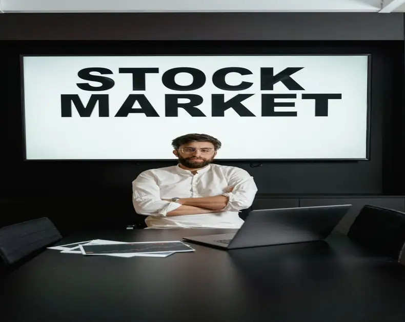 Man in a white shirt stands with arms folded in a dark conference room, a large 'STOCK MARKET' sign on screen behind him with a laptop and papers on the table.