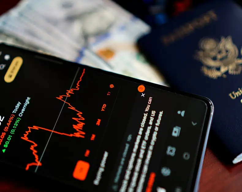 Smartphone screen showing a red line graph against a dark UI, with cash and a passport blurred in the background, suggesting travel or financial activity.